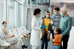 female doctor in a white lab coat and blue gloves speaking with a multi-generational family wearing face masks in a medical clinic waiting room