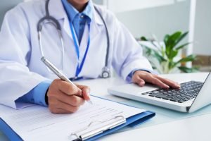 A close-up of a female medical professional in a white lab coat with a stethoscope around her neck, filling out a form on a clipboard with her right hand while working on a laptop with her left hand