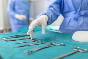Nurse hand taking surgical instrument for group of surgeons at background operating patient in surgical theatre. Steel medical instruments ready to be used.