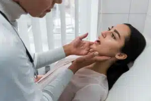 A doctor wearing coat and stethoscope checking the throat of a female patient lying on a bed in urgent care facility