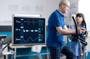 An elderly man undergoing a cardiac stress test on an elliptical machine while a medical professional monitors his vital signs on a screen displaying heart rate graphs