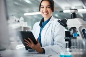A smiling female medical professional or researcher in a white lab coat holding a tablet next to a microscope in a laboratory setting