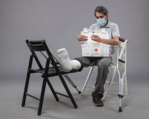 A man wearing a face mask sits with his casted leg propped on a chair, holding a first aid kit next to a crutch