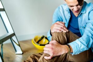 A man sitting on a floor holding his injured leg while his safety helmet is placed beside him