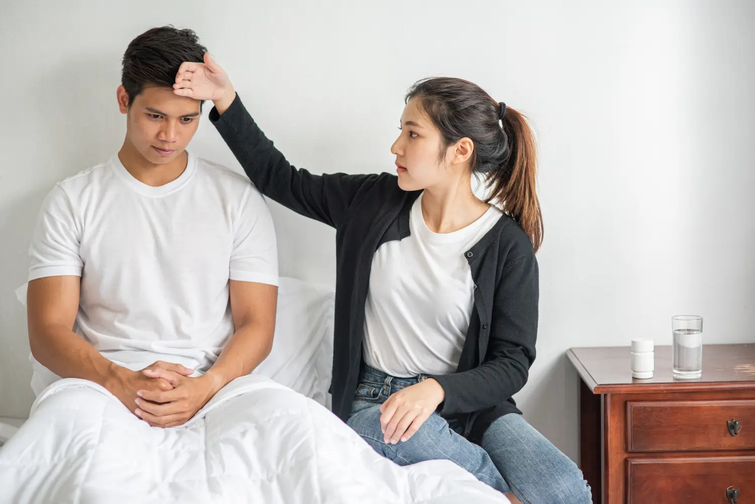 A concerned young woman places her hand on a man's forehead while he sits up in bed looking unwell.