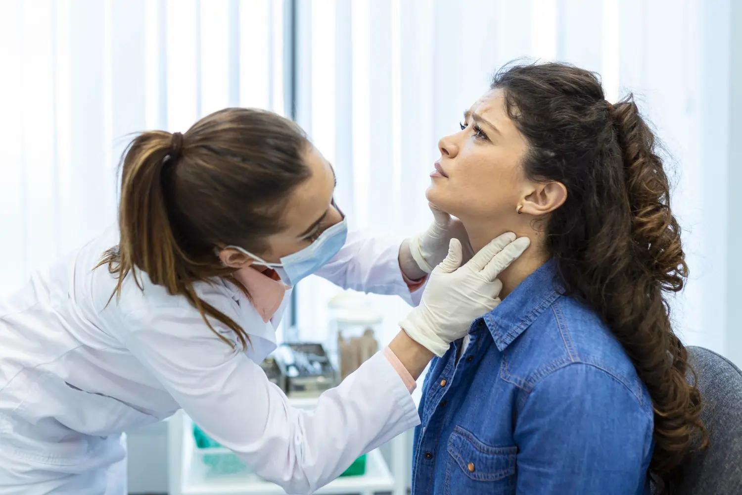A female healthcare professional wearing a face mask and medical gloves carefully palpates the neck of a concerned young woman during a physical examination.