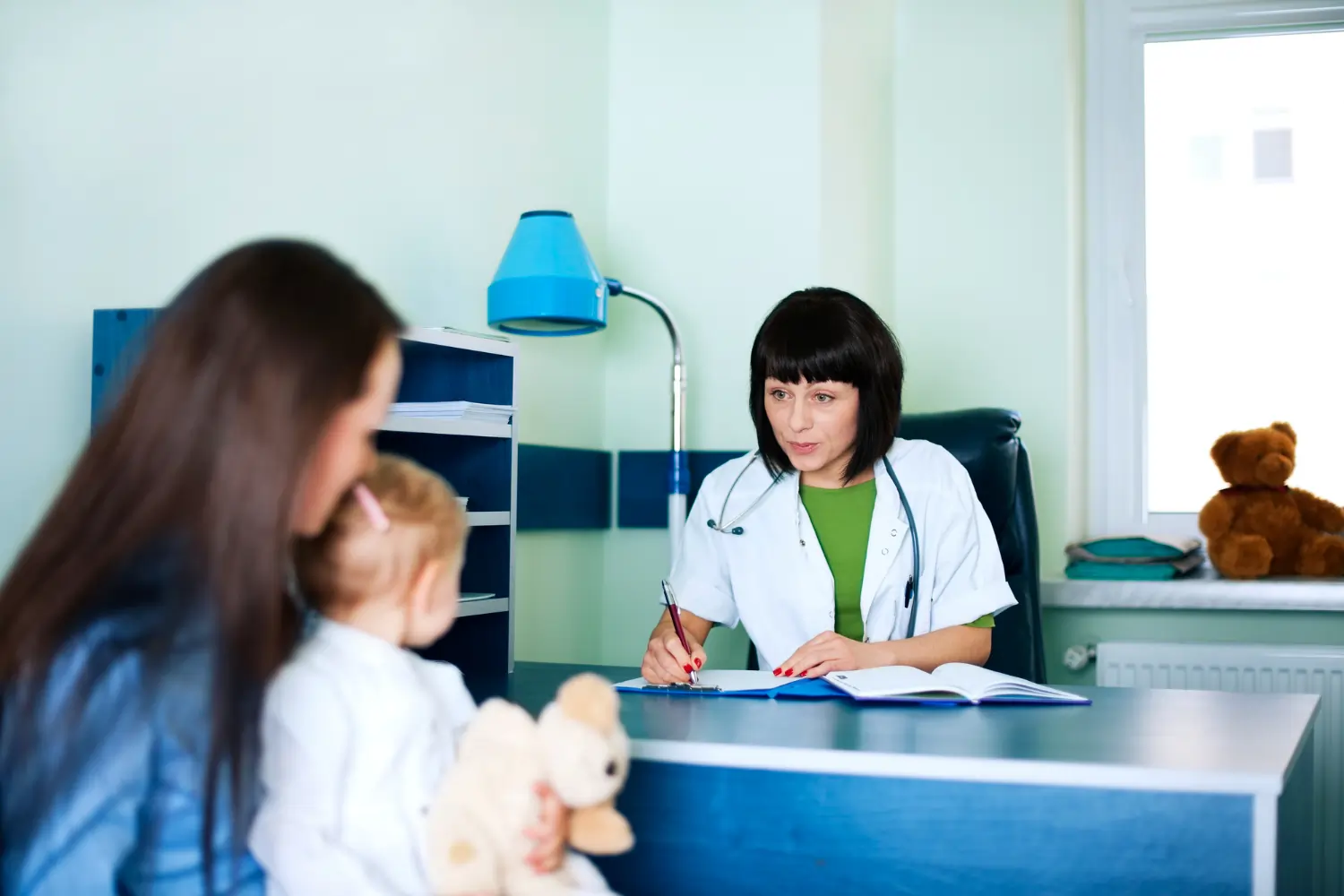 A mother sits with her toddler daughter holding a teddy bear, consulting a female pediatrician who is writing on a clipboard at a blue clinic desk next to a window.