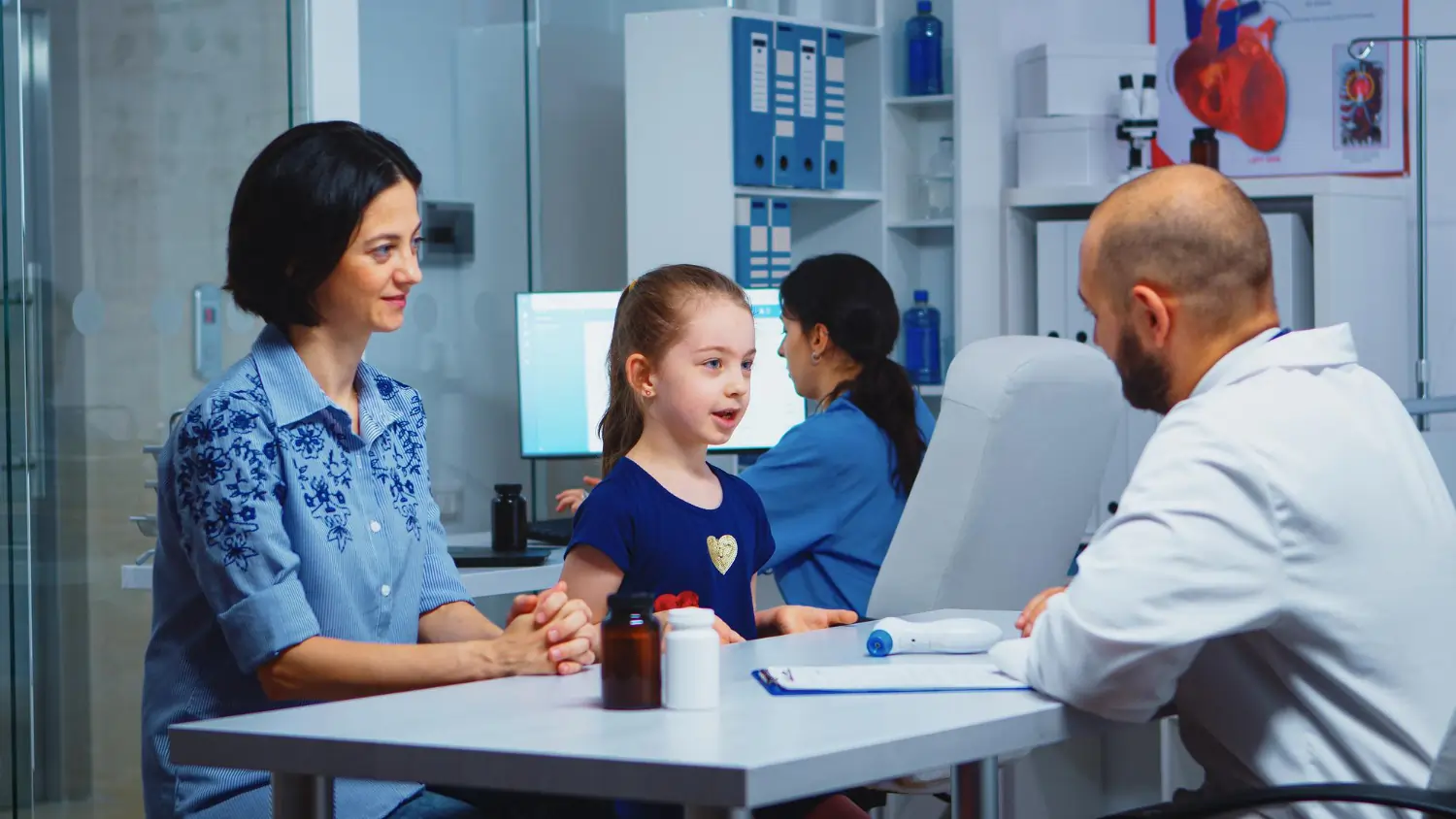 A young girl speaks with a male pediatrician sitting across a clinic desk, while her mother sits beside her listening carefully. A medical assistant works on a computer in the background next to a cardiovascular anatomy poster.