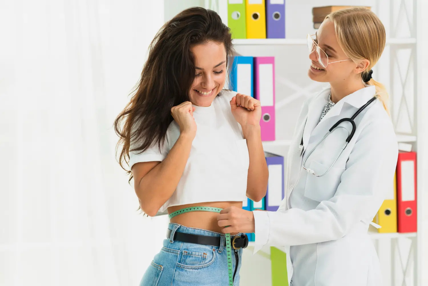 A cheerful female doctor or nutritionist in a white coat wraps a green measuring tape around a young woman's waist, while the patient smiles excitedly with raised fists during a weight loss checkup.